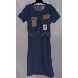 May include: A blue denim dress featuring a short-sleeved bodice with a button-down front, a round neckline, and a long skirt. The bodice includes rectangular patches with embroidered red floral designs and a plain brown patch.