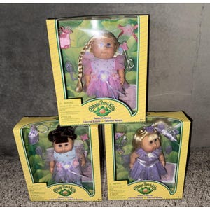 Puede incluir: Tres muñecas Cabbage Patch Kids de la colección Fantasía en sus cajas originales. Cada muñeca tiene un peinado único y viste un vestido brillante. Las cajas son amarillas con detalles verdes.