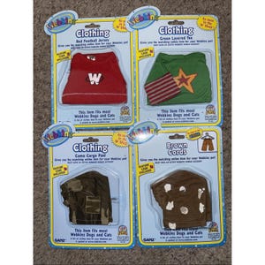 Puede incluir: Cuatro prendas de vestir Webkinz en embalaje. Incluye una camiseta de fútbol roja con una "W" blanca, una camiseta verde en capas con una estrella y rayas, pantalones cargo de camuflaje y cordones marrones. Cada paquete indica que el artículo se adapta a Webkinz Dogs and Cats.