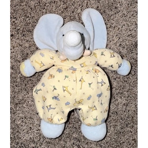 May include: A plush elephant toy with light blue ears, trunk, and feet. The elephant wears a yellow outfit patterned with various animal and object designs. The toy is designed for babies and young children.