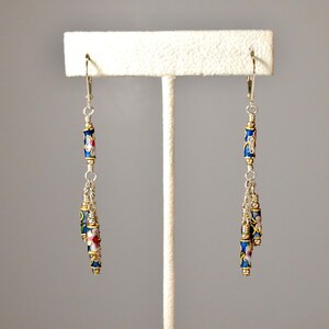 May include: A pair of dangle earrings with blue and white enamel beads and gold accents. The earrings are hanging from silver hooks.