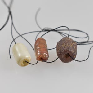 May include: Three beads on black string. One is off-white and cylindrical, the second is reddish-orange with etched lines, and the third is a dark brown, hexagonal shape. The beads are strung on black thread.