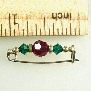 May include: A silver safety pin with a red and green beaded design. The pin is approximately 1 inch long.