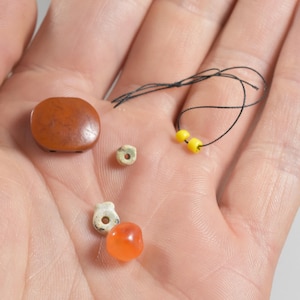 Könnte beinhalten: Eine Sammlung kleiner Perlen und ein Knopf in verschiedenen Formen und Farben. Der Knopf ist ein abgerundetes Quadrat, und die Perlen sind rund und zylindrisch. Die Farben umfassen Braun, Orange, Gelb und Off-White. Ein schwarzer Faden ist ebenfalls vorhanden.