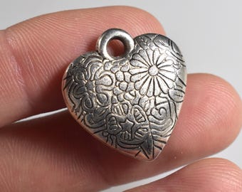 Könnte beinhalten: Silberner herzförmiger Anhänger mit aufwendigen Blumen- und Wirbelmustern. Der Anhänger hat oben eine Schlaufe zum Befestigen an einer Kette oder einem Band. Das Design ist detailliert und geprägt, wodurch eine strukturierte Oberfläche entsteht.