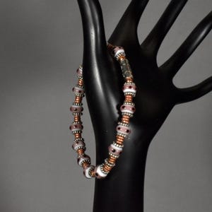 Könnte beinhalten: Ein Armband mit weißen und malvenfarbenen Perlen mit schwarzen Punkten, kupferfarbenen Abstandshaltern und silberfarbenen Metallakzenten. Das Armband wird auf einem schwarzen, handförmigen Display präsentiert.