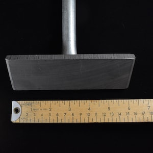 Op de afbeelding: Een rechthoekig, donkergrijs metalen gereedschap met een handvat. Een houten liniaal toont inches en centimeters. Het rechthoekige deel van het gereedschap is ongeveer 15 cm lang.