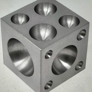 Könnte beinhalten: Ein silberner Metallwürfel mit unterschiedlich großen konkaven Vertiefungen und Löchern. Der Würfel ist ein Werkzeug zur Schmuckherstellung, das zum Formen und Formen von Metall bestimmt ist. Der Würfel hat eine strukturierte Oberfläche.