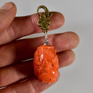 Könnte beinhalten: Ein orangefarbener, ovaler Anhänger mit detaillierten Schnitzereien von Vögeln und Blattwerk. Der Anhänger ist an einem messingfarbenen Karabinerverschluss in Form einer Blume mit einem Blatt befestigt, verbunden durch eine silberfarbene Drahtwicklung.