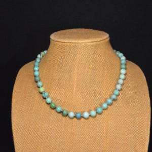Op de afbeelding: Een turquoise kralenketting tentoongesteld op een bruine etalagepop. De ketting heeft ronde, lichtblauwe en groene kralen die aan elkaar zijn geregen. De kralen hebben een diameter van ongeveer 0,8 cm. De achtergrond is zwart.