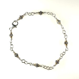 Op de afbeelding: Een zilveren kettingarmband met gouden accenten en kleine ronde kralen. De armband heeft een karabijn sluiting.