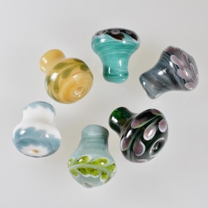 Op de afbeelding: Een verzameling van acht glazen flessenstoppers met verschillende kleuren en patronen. De stoppers zijn gevormd als paddenstoelen en hebben ontwerpen met bladeren, swirls en stippen.