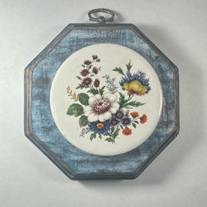 May include: A decorative wall hanging featuring a round, floral-patterned ceramic tile set within a distressed, octagonal blue wooden frame. The floral design includes white, purple, yellow, and orange blooms with green foliage. A metal loop is at the top for hanging.