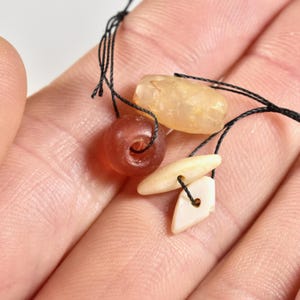 May include: Close-up of assorted beads on black string. The beads include a translucent amber-colored oblong bead, a reddish-brown round bead, and two off-white, irregular-shaped beads. The beads are displayed against a light background.