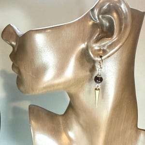 Könnte beinhalten: Ein einzelner Ohrring mit einem silbernen Haken, einer dunkelroten Perle und einem goldfarbenen Kegelanhänger. Der Ohrring wird auf einem Schaufensterkopf präsentiert. Ein einzigartiger Schmuck.