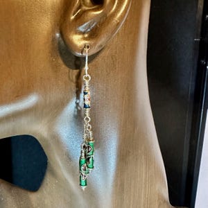 Könnte beinhalten: Hängeohrring aus Silber mit Haken und Kette. Er hat eine kleine, zylindrische Perle mit floralem Design, gefolgt von drei grünen, zylindrischen Perlen mit aufwendigen Silberdetails. Der Ohrring ist für das Tragen in einem durchbohrten Ohr konzipiert.