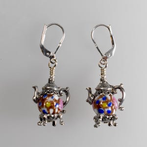 May include: A pair of silver-toned teapot earrings. The teapots have a colorful enamel design with white, brown, yellow, blue, and pink floral patterns. The earrings have lever-back closures.