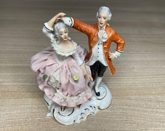 May include: A porcelain figurine of a couple dancing. The woman is wearing a pink dress with lace and the man is wearing a red and white jacket with black pants. The figurine is on a white base with gold accents.