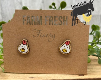 May include: A pair of wooden chicken earrings with red combs, white bodies, and yellow beaks. The earrings are displayed on a brown card with the text "Farm Fresh Finery".