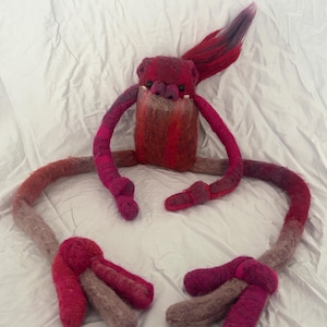 May include: A needle-felted creature doll featuring a rectangular body in shades of magenta, red, and dark pink. It has long, thin, flexible arms and legs ending in large, three-toed feet. The head has two small black eyes and a tuft of red hair.