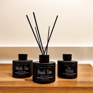 May include: Three black reed diffusers on a wooden surface. The center diffuser has black reeds inserted. Each bottle has a label with white text. The labels read "White Tea", "Black Sea", and "Sea Minerals". Each bottle is 3.5 oz net weight.