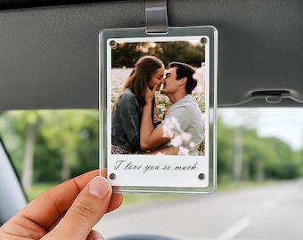 May include: Clear acrylic car charm with a photo of a couple kissing in a field. The charm is rectangular and has the text "I love you so much" in script. The charm is hanging from a car visor.