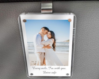 May include: Clear acrylic car visor photo holder with a photo of a couple embracing on a beach. The photo holder is attached to a gray car visor with a metal clip. Text on the photo reads, "Every mile, I'm with you. Drive safe."