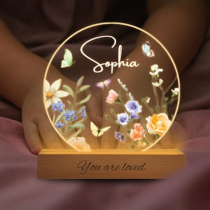 May include: A personalized night light with the name "Sophia" in elegant script. The circular design features colorful flowers and butterflies, illuminated by a warm glow. The wooden base reads "You are loved".