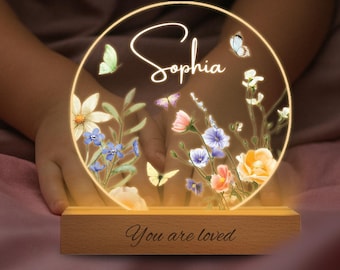 May include: A personalized night light with the name "Sophia" in elegant script. The circular design features colorful flowers and butterflies, illuminated by a warm glow. The wooden base reads "You are loved".