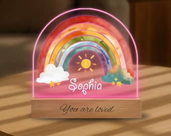 May include: A rainbow-shaped night light with a wooden base. The rainbow features vibrant colors and a sun graphic. The name "Sophia" is written across the front, with the phrase "You are loved" below.