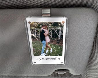 May include: Clear acrylic car visor photo clip with a photo of a couple embracing. The photo clip is attached to a gray car visor. The text "My entire world <3" is printed on the photo.