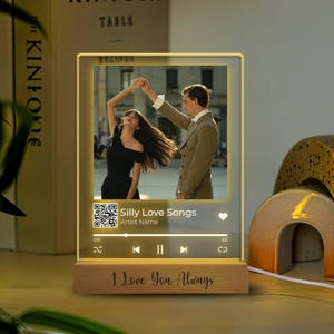 May include: A wooden photo frame with a lit up screen displaying a couple dancing. The screen also shows a music player with the song title "Silly Love Songs" and the artist name. The frame has a wooden base with the text "I Love You Always" engraved on it.