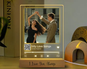 May include: A wooden photo frame with a lit up screen displaying a couple dancing. The screen also shows a music player with the song title "Silly Love Songs" and the artist name. The frame has a wooden base with the text "I Love You Always" engraved on it.