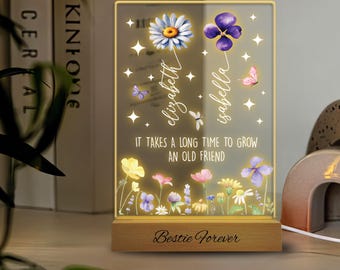 May include: A glass light-up sign with a floral design and the text "It takes a long time to grow an old friend." The sign also has two names, "elizabeth" and "isabella", written vertically on the left and right sides of the floral design. The sign is on a wooden base with the text "Bestie Forever" engraved on it.