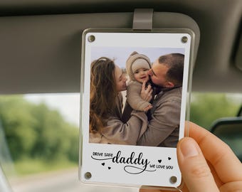 May include: Clear acrylic car visor photo holder with a family photo. The photo shows a family of three. The text on the holder reads "Drive Safe Daddy We Love You."
