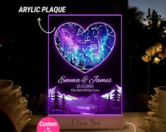 May include: A personalized acrylic plaque with a heart-shaped constellation design. The plaque features the names "Emma & James" and the date "12.12.2021" with the text "The Start Of Our Love". The plaque is illuminated with purple light and sits on a wooden stand with the text "I Love You".