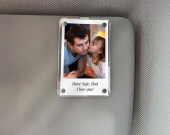 May include: A clear acrylic photo holder with a picture of a man and a child kissing. The text "Drive Safe, Dad I love you!" is printed at the bottom. The holder is clipped to a gray car visor.