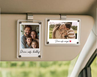May include: Two clear acrylic photo holders attached to a car visor. Each holder contains a family photo with the text "Drive safe, daddy!" and "Drive safe, amigos! ❤️". The photos are held in place with silver metal clips.