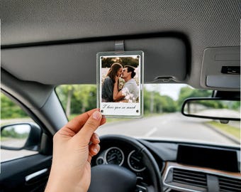 May include: Clear acrylic car charm with a photo of a couple kissing in a field. The charm is rectangular and has the text "I love you so much" in script. The charm is hanging from a car visor.