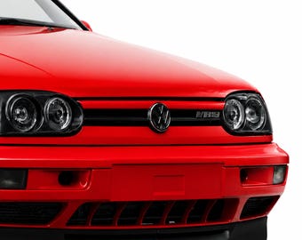 May include: The front end of a bright red Volkswagen Golf vehicle featuring black dual-lens headlights, a central black Volkswagen emblem, and a VR6 badge on the grille. The bumper includes a lower black air intake vent and a black front lip spoiler.
