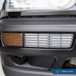 May include: A black car bumper featuring a clear headlight lens at the top, an amber turn signal light, and a rectangular white fog light with horizontal slats. The bumper includes a lower air intake vent and the text GOLF and PROJECT PARTS.