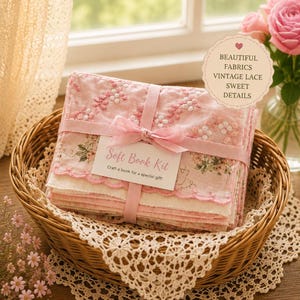 May include: A soft book kit featuring stacked fabric pages with pink floral embroidery and scalloped edges, tied with a pink ribbon. A white tag reads Soft Book Kit, Craft a book for a special gift, and a circular label says BEAUTIFUL FABRICS VINTAGE LACE SWEET DETAILS.