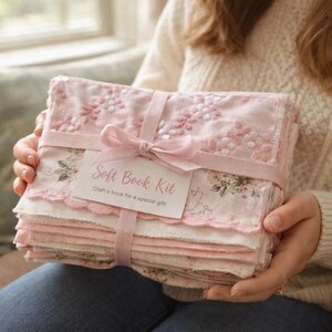 May include: A stack of fabric pages for a soft book kit, tied with a pink ribbon. The top page features pink floral embroidery and scalloped edges. A white tag attached to the ribbon reads Soft Book Kit, Craft a book for a special gift.