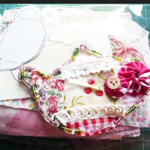 May include: A handmade fabric teapot applique with floral and cherry patterns, adorned with lace and a button. The applique is embellished with beads and sits atop a stack of pink and floral fabric pieces, with a paper template visible.