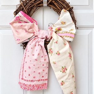 May include: A natural brown twig wreath adorned with a large, tied fabric bow. The bow is constructed from two distinct cotton fabrics: one side features a light pink background with small floral prints, and the other side is cream with pink and green floral patterns.