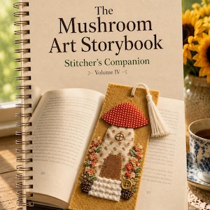 Può includere: Un libro intitolato "The Mushroom Art Storybook" con un segnalibro in feltro con un design a forma di casa a fungo. Il segnalibro ha un cappello a fungo a pois rossi e bianchi e un nappa bianca. Una tazza di tè e girasoli sono sullo sfondo.