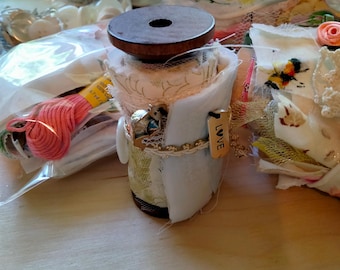 May include: A decorative spool with a wooden top and a white, fabric-covered body. The spool is adorned with lace, beads, and a small tag that reads "LOVE". Pink embroidery floss and other craft supplies are in the background.