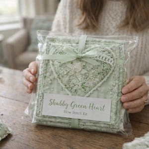 May include: A "Shabby Green Heart" slow stitch kit, packaged in clear plastic. The kit contains green fabric squares, decorated with a heart-shaped design. A light green ribbon and a button are visible. The kit is presented on a wooden table.