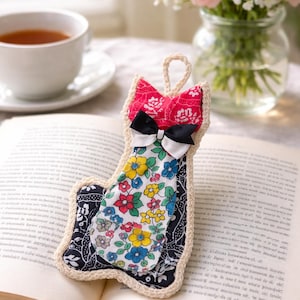 May include: A handmade fabric bookmark with a floral pattern, a black and white bow, and a red top. The bookmark is edged with cream-colored crochet and is resting on an open book.