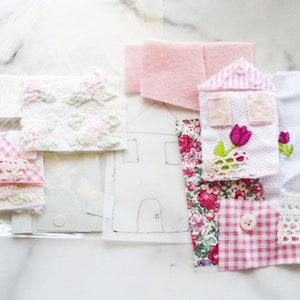 Könnte beinhalten: Eine Sammlung von Stoffresten in Rosa, Weiß und Blumenmustern, darunter ein kleines hausförmiges Stück mit Stickereien. Die Stoffe sind auf einer weißen Oberfläche angeordnet.